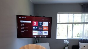 A living room with a flat-screen TV mounted on the wall displaying a streaming service interface. A remote control is on a table, and a fan is on the table. - Modern condo near Microsoft for business and leisure travelers (Bellevue)