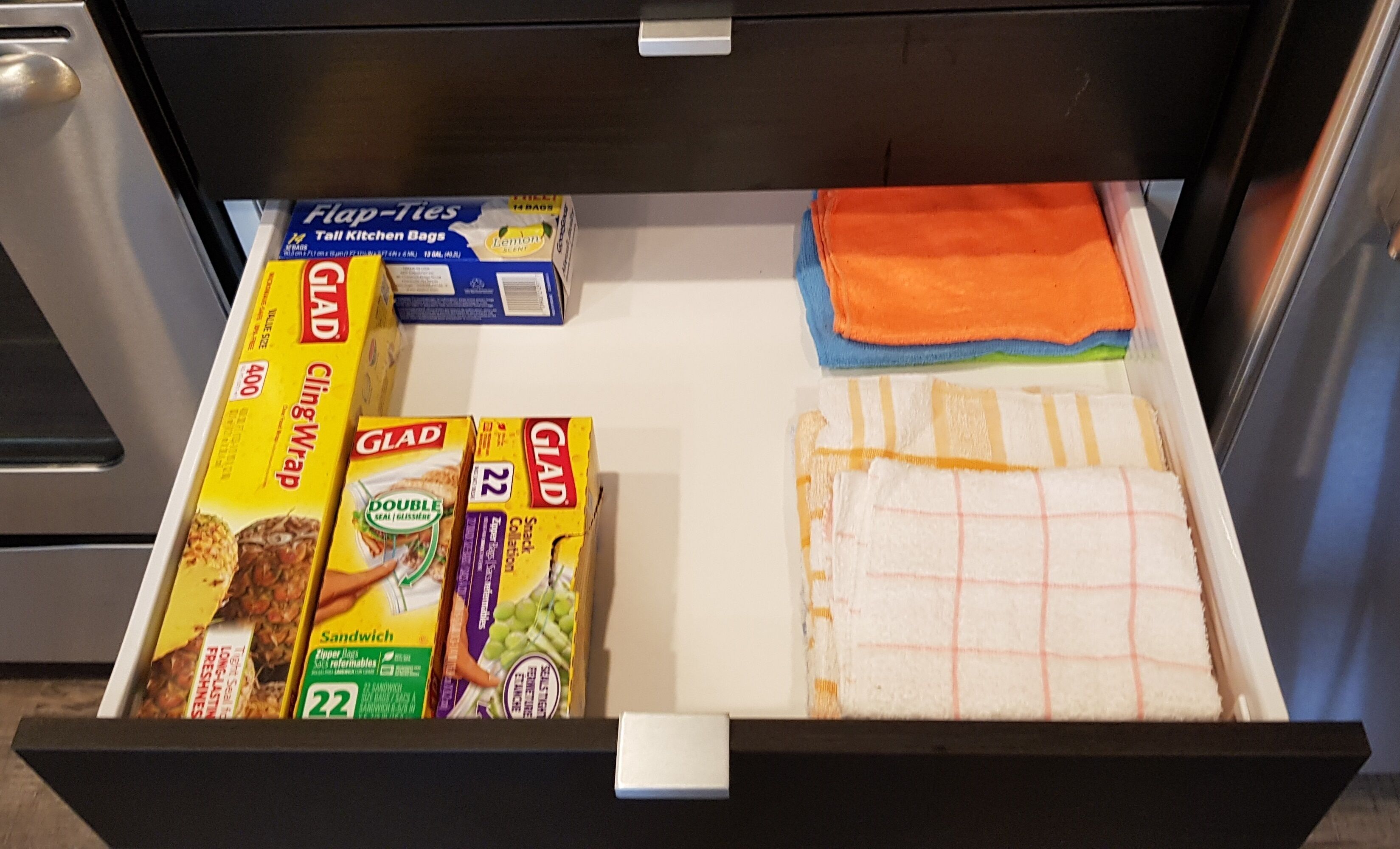 Un tiroir contenant des articles de rangement pour la cuisine, notamment des sacs à fermeture éclair pour sandwichs, du film alimentaire et un torchon à motifs quadrillés.