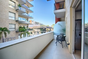 A balcony with a view of a building with multiple floors and balconies, a small table with two chairs, and a refrigerator inside.