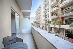 En balkon med stol og bord, ud til en gade med bygninger og palmer.