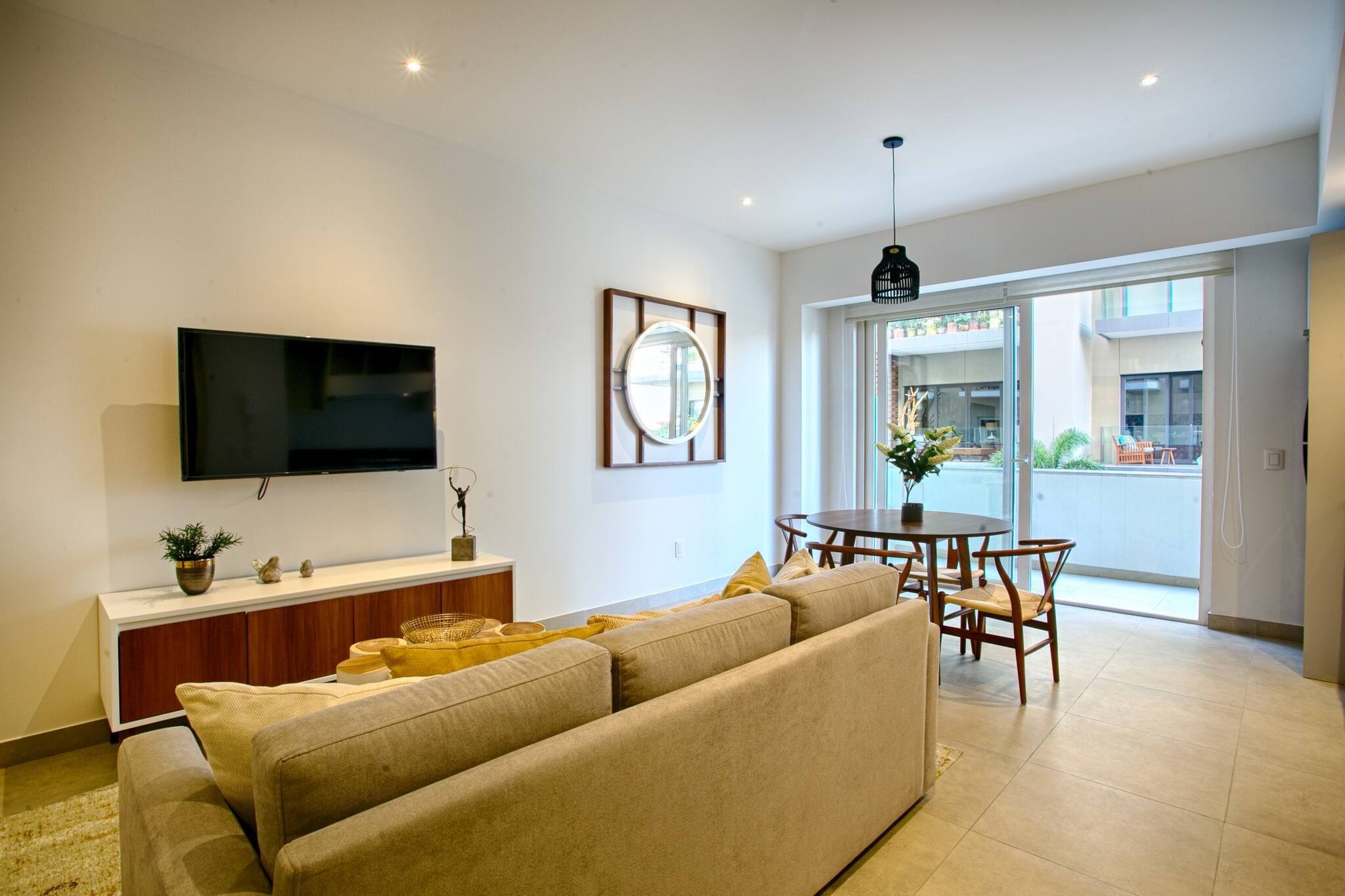 En moderne stue med et fladskærms-tv, et rundt spejl, en sofa, et spisebord med stole og en balkon med udsigt.