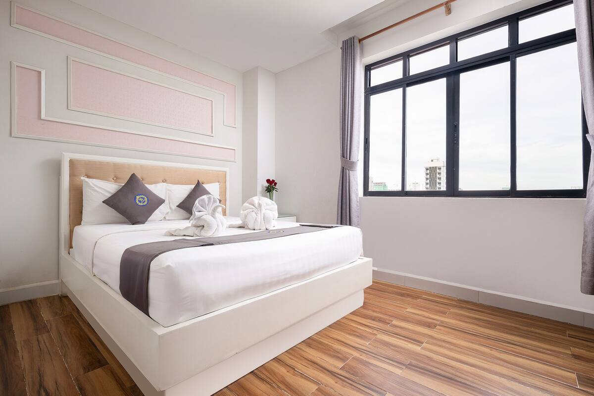 Ein ordentlich bezogenes Bett mit Holz-Kopfstück, ein großes Fenster mit Vorhängen und ein Holzboden.