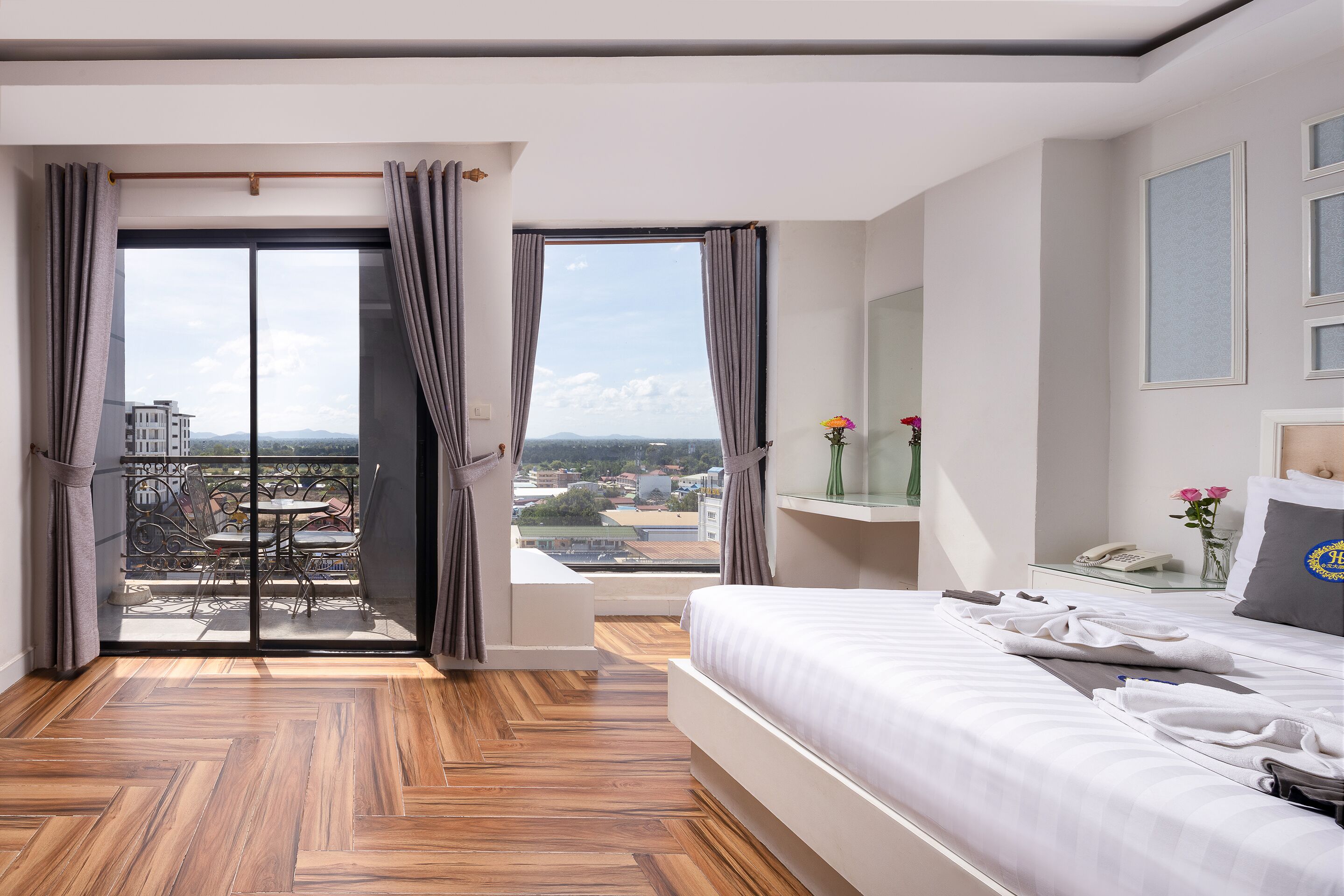 A hotel room with a large bed, a bedside table with a phone and a vase of flowers, a view of the city through a sliding door, and a wooden floor.
