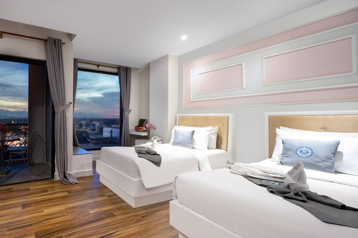 Ein Hotelzimmer mit zwei Betten, einem Balkon mit Blick und einer Stadtansicht durch das Fenster.