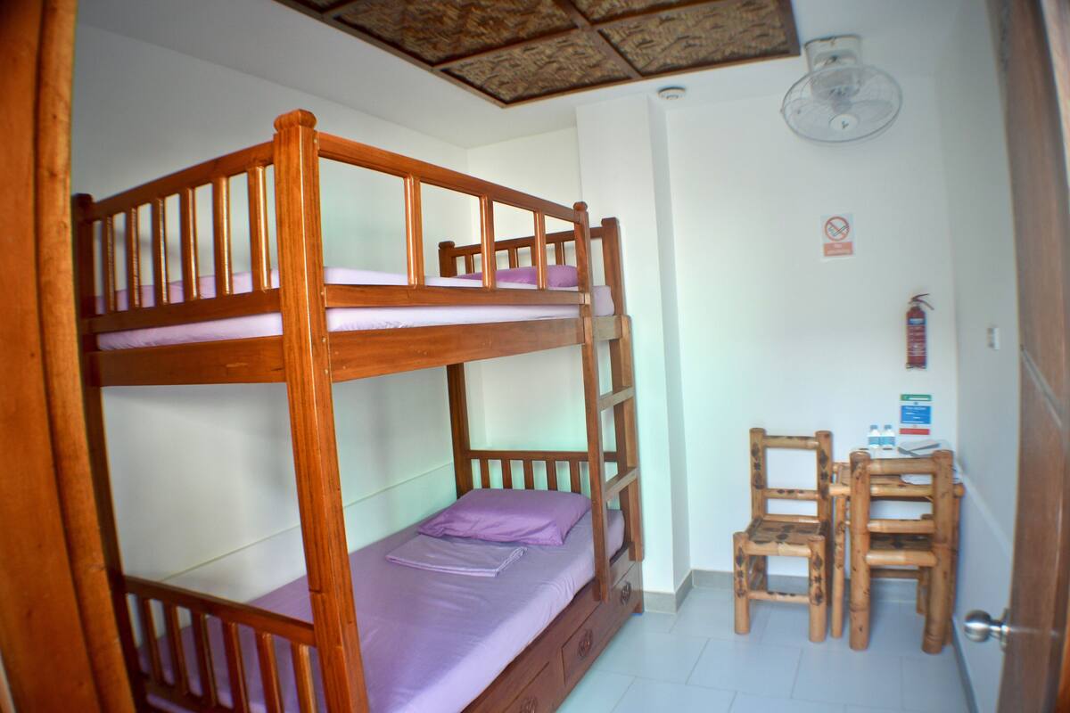 A bunk bed with a wooden frame and purple bedding, a wooden chair, and a small table with a water bottle and a notepad.