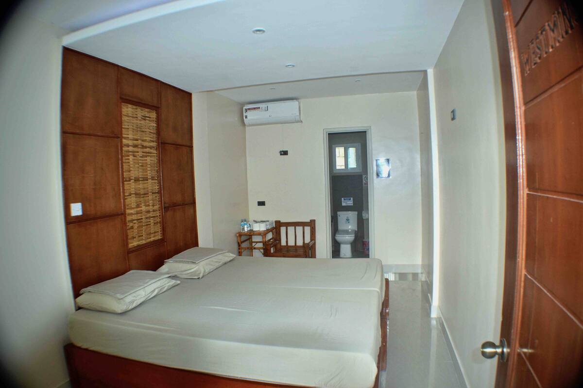 A bedroom with two beds, a wooden wardrobe, a small table with a chair, and an air conditioning unit on the wall.