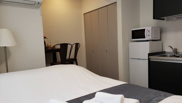 A hotel room with a bed, a desk with a chair, a kitchenette, and a wardrobe. - Ocean View Araha 201 (Chatan)