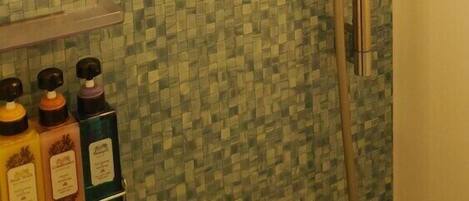 A bathroom with mosaic tile walls, a shower with a handheld showerhead, and a shelf with toiletries.
