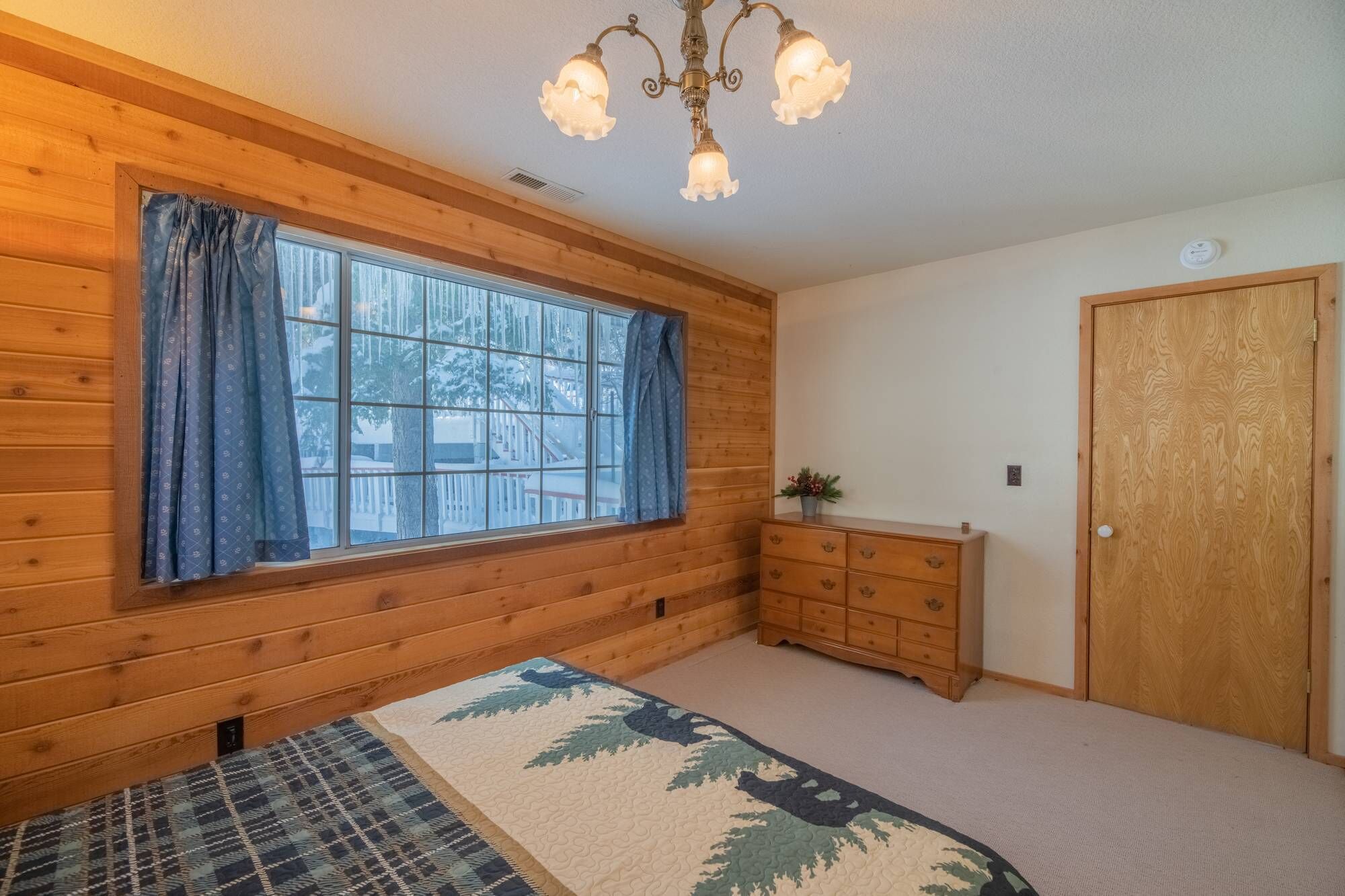 Un dormitorio con paredes de madera, un ventanal con cortinas, una cama con cubrecama estampado, un aparador y una puerta.