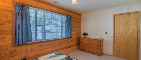 Un dormitorio con paredes de madera, un ventanal con cortinas, una cama con cubrecama estampado, un aparador y una puerta.