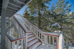 Una escalera con barandillas blancas y detalles rojos que conduce a una zona cubierta, con árboles nevados al fondo.