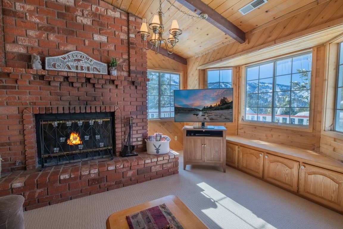 Interior de una cabaña con chimenea de ladrillo, vigas de madera, televisor y vistas al exterior.