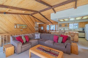 A cabin interior with a wooden ceiling, a sofa with cushions, a coffee table, and a kitchen area.