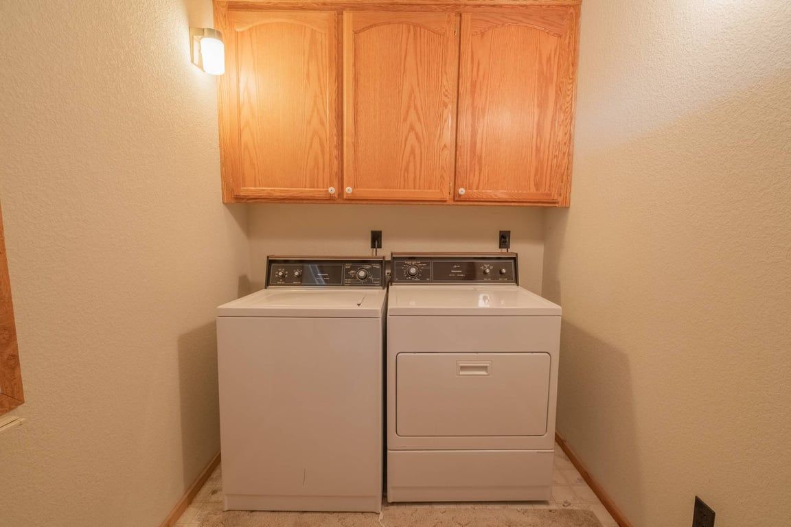 A laundry room with two stacked washers and dryers, wooden cabinets above, and a light fixture on the left wall.