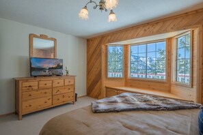 Ein Schlafzimmer mit einer Holzwand, einem großen Fenster mit Blick auf Bäume, einem Bett mit Felldecke und einer Holzkommode mit Fernseher.