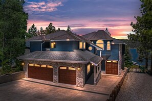 Une maison à deux étages avec une façade en pierre, deux portes de garage et une vue sur un lac au coucher du soleil.