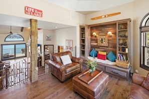 A living room with leather furniture, a stone pillar, a wooden bookshelf, and a sign that reads 'LIFE IS BETTER AT THE LAKE'. - Over 5,700 Square Feet of Lakeside Luxury! Private Dock! Hot Tub! Game Room! Arcade Room! Beach! Patio Gazebo! Ornate Iron and Marble Accents! (Big Bear Lake)