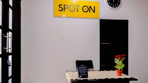 Una mesa de recepción con un letrero amarillo que dice "SPOT ON", un reloj y una planta en maceta con flores rojas.