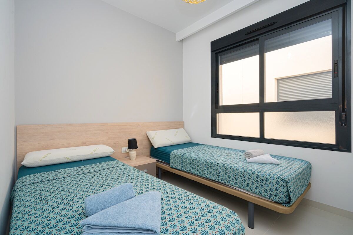 A room with two beds, each with patterned bedding, a bedside table with a lamp, and a window with blinds.