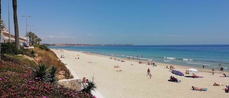 A beach with people sunbathing and swimming, palm trees, and a clear blue sky.