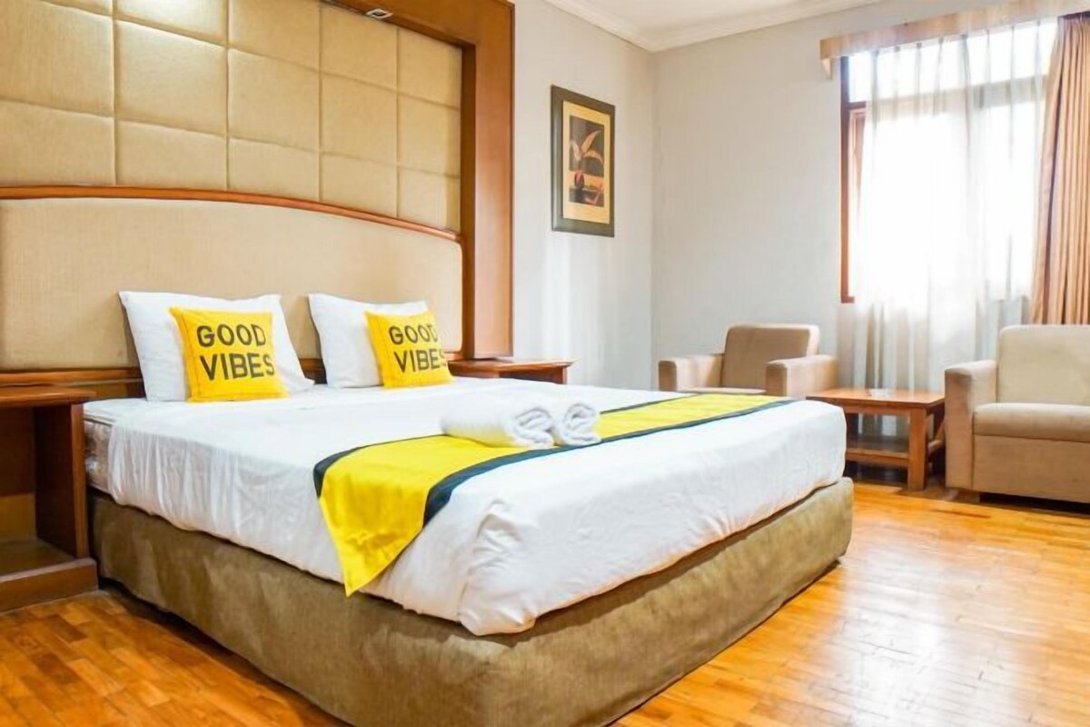 A hotel room with a large bed, two pillows with 'GOOD VIBES' text, a wooden headboard, a wooden floor, a small table, and a chair.