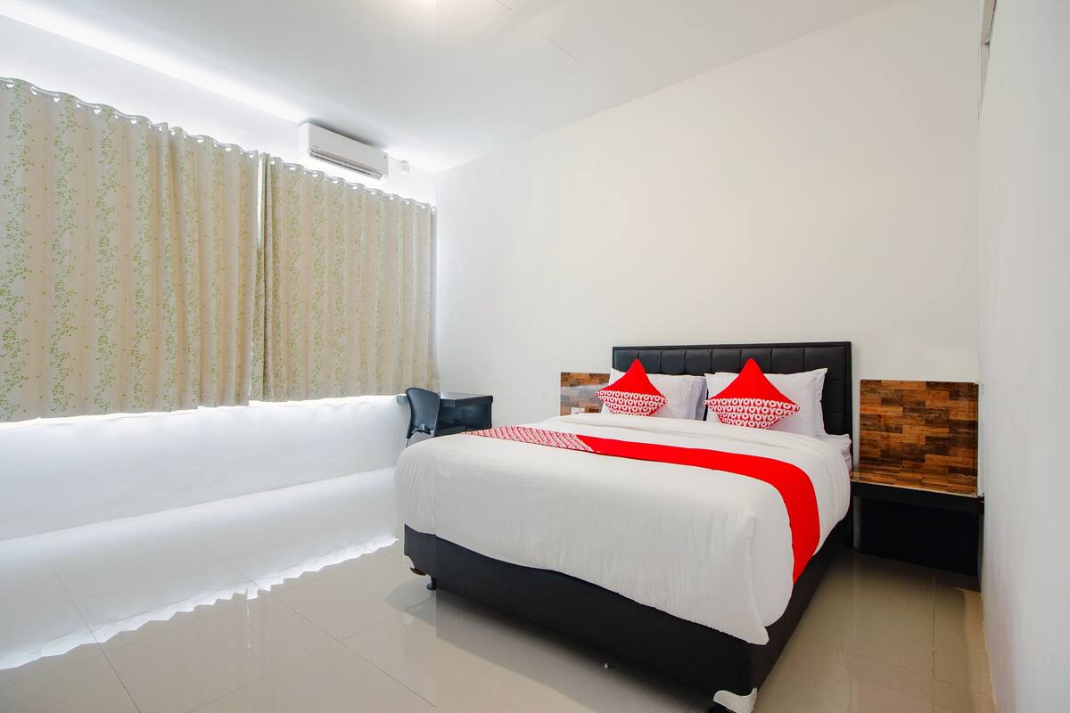 A modern hotel room with a large bed, a wooden headboard, a nightstand, and a window with curtains.