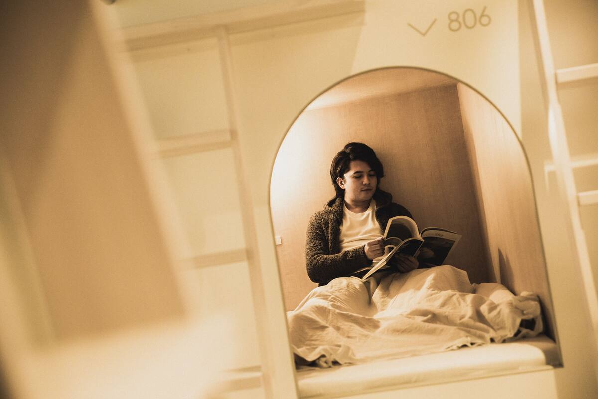 Une personne est assise sur un lit, un livre à la main, dans une pièce où l’on peut voir le numéro « 806 » sur le mur.