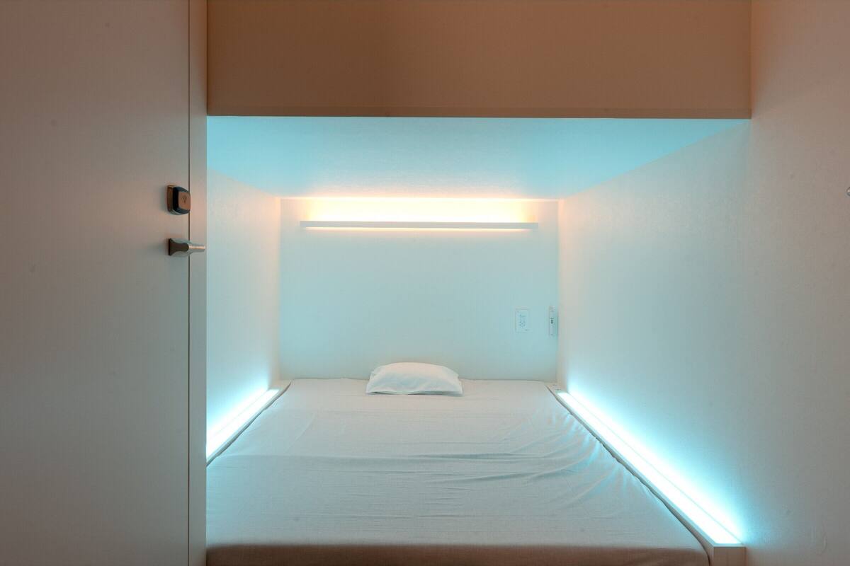 Une petite pièce bien éclairée, avec un lit simple, une table de chevet et un luminaire fixé au mur.