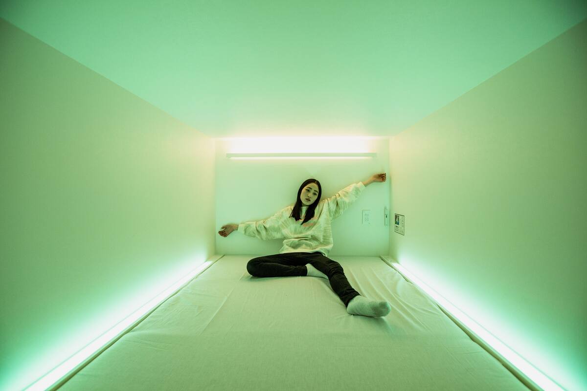 Une personne est assise sur un lit dans une pièce aux nuances verdâtres, éclairée par un luminaire fixé au plafond.