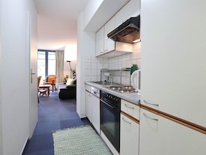 Una cocina estrecha con armarios blancos, una estufa y un lavamanos. Se ve un rincón comedor con una mesa y sillas al fondo.