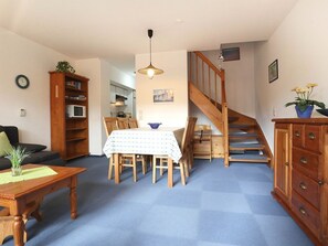 Una sala de estar con una mesa de comedor, sillas, un sofá, una estantería, una escalera y una cocina al fondo.