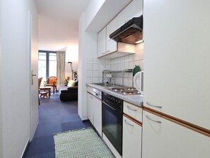 Een smalle keuken met witte kasten, een fornuis en een spoelbak. In de achtergrond is een eethoek met een tafel en stoelen te zien.
