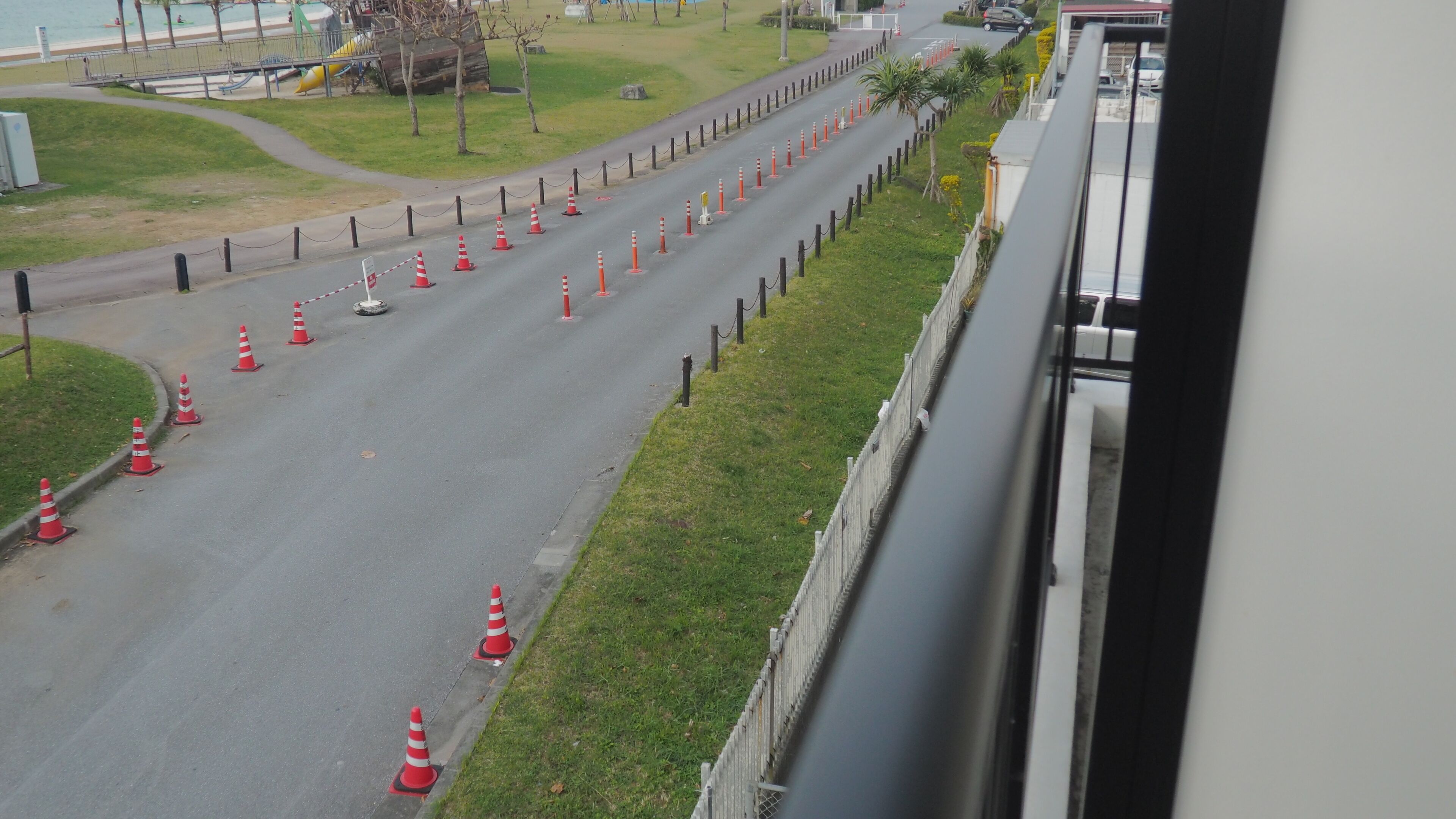 A road lined with traffic cones and a fence, with a grassy area and a building in the background.