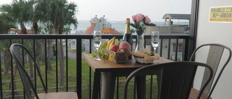 Een balkon met een tafel gedekt voor twee personen, met een fles champagne, een vaas met roze bloemen en een fruitmand.