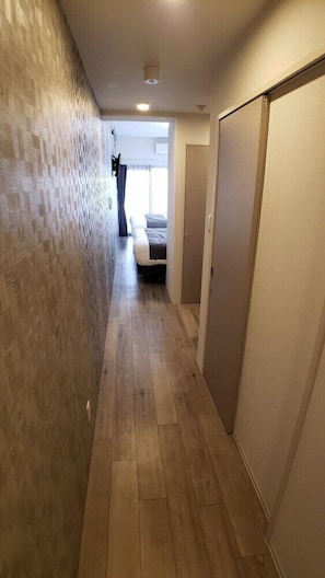 A narrow hallway with wooden flooring, a textured wall on one side, and a bed with a gray bedspread in the background.