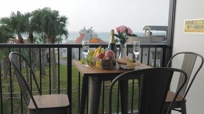 A balcony with a table set for two, featuring a bottle of champagne, a vase with a pink flower, and a basket of fruit. - Ocean View Araha 205 (Chatan)