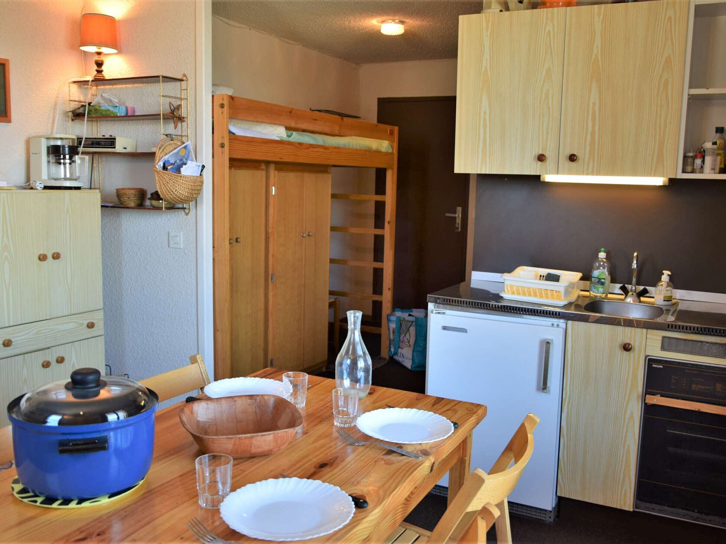 Une cuisine compacte avec une table en bois dressée pour le repas, un petit réfrigérateur et une machine à café posée sur une étagère.