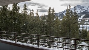 A balcony with a metal railing overlooking a snowy mountain landscape with trees and a building in the background. - Community Sauna | Mountain Views (Big Sky)