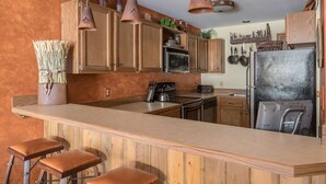 A kitchen with wooden cabinets, a central island with stools, a refrigerator, and hanging pots. - Community Sauna | Mountain Views (Big Sky)