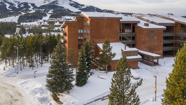 A multi-story building with a brick facade, surrounded by snow-covered ground and trees, with a mountain in the background. - Community Sauna | Mountain Views (Big Sky)