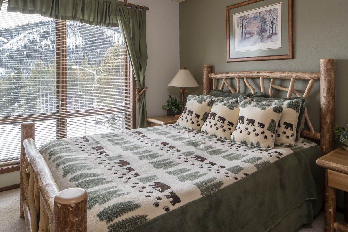 A cozy bedroom with a wooden headboard, a large window with curtains, a bed with a green and white patterned blanket, and a framed picture on the wall.