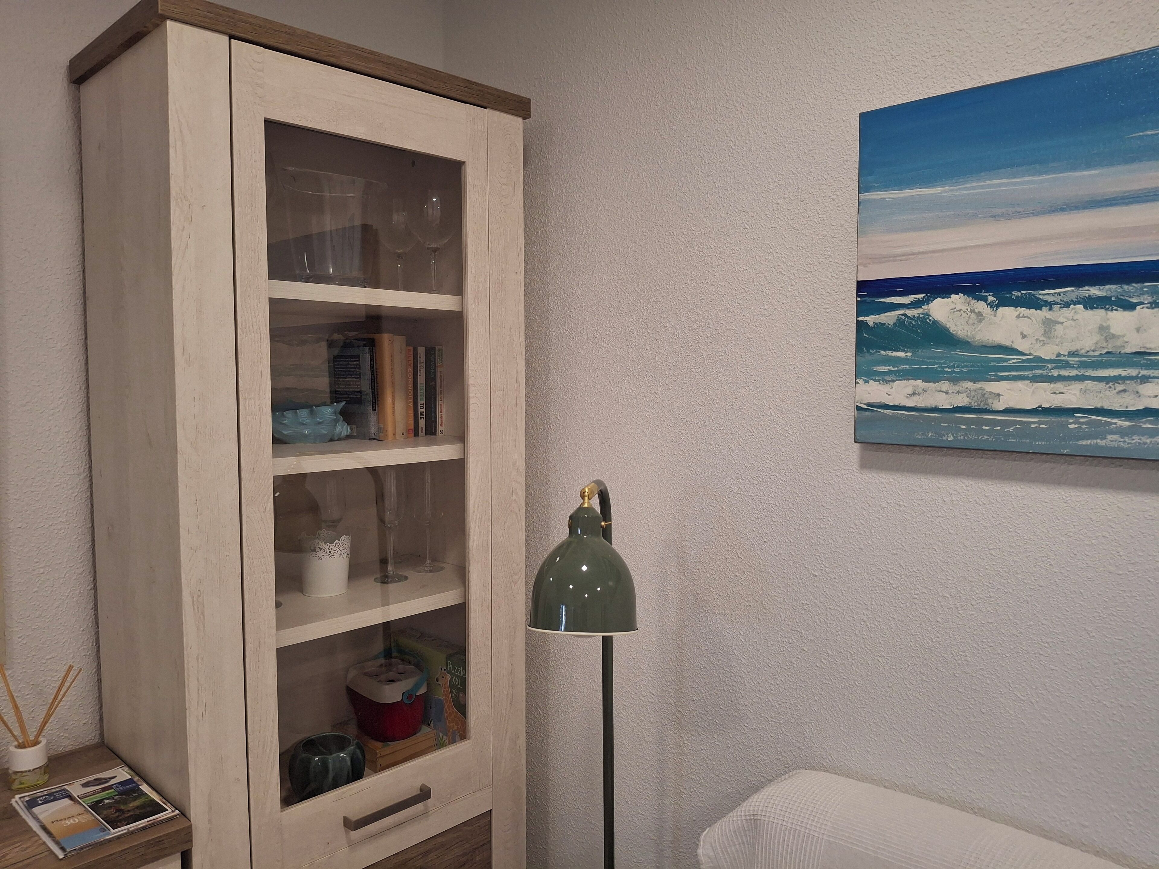 A room with a wooden cabinet, a floor lamp, a painting of the ocean, and a small table with a magazine and a vase.