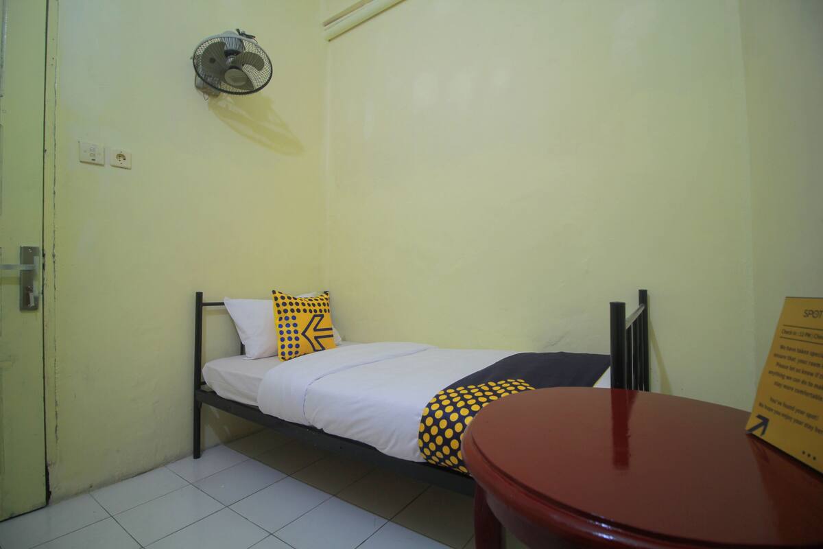 Een kleine eenpersoonskamer met een bed, een tafel en een ventilator aan de muur.