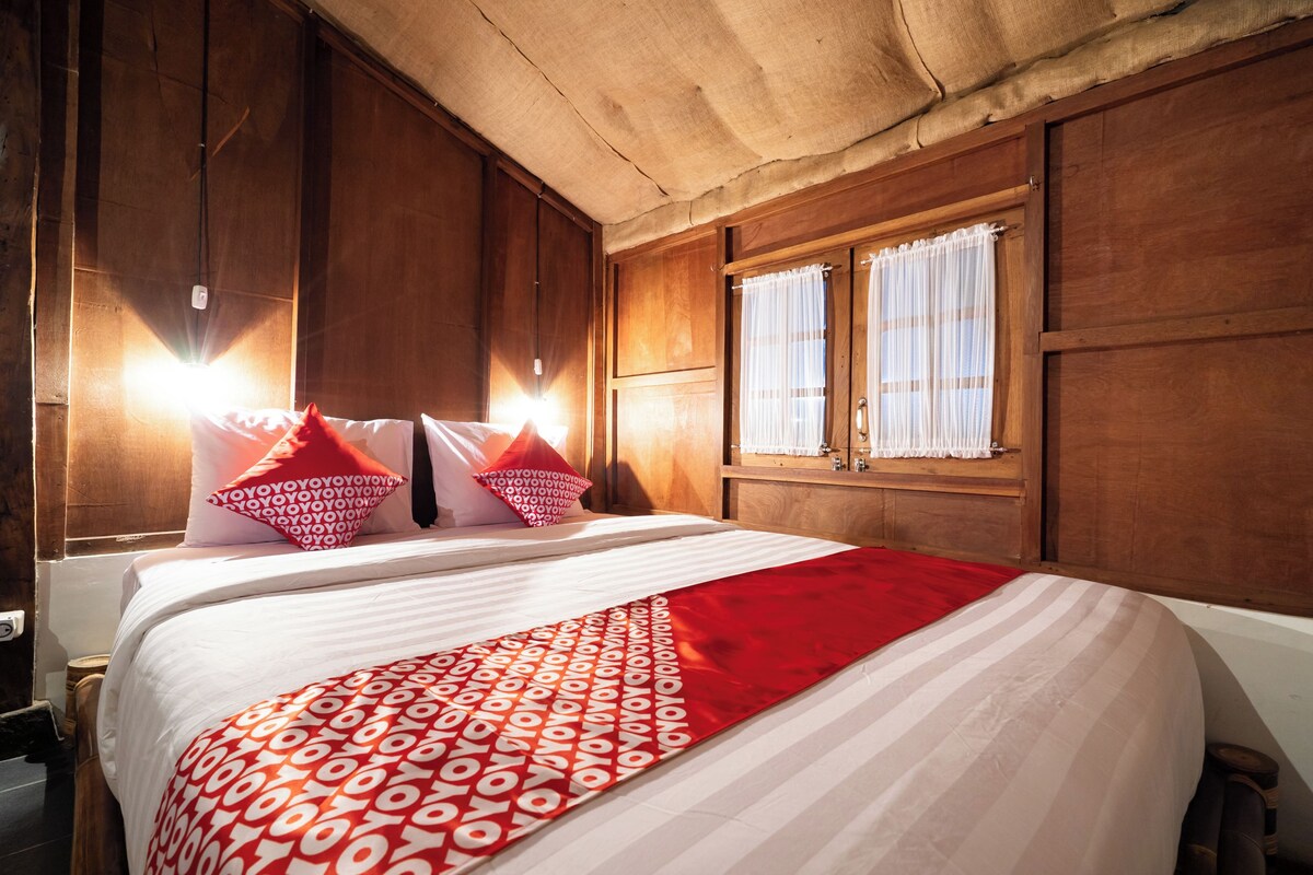 A neatly made bed with a red and white patterned bedspread, wooden walls, and a window with sheer curtains.