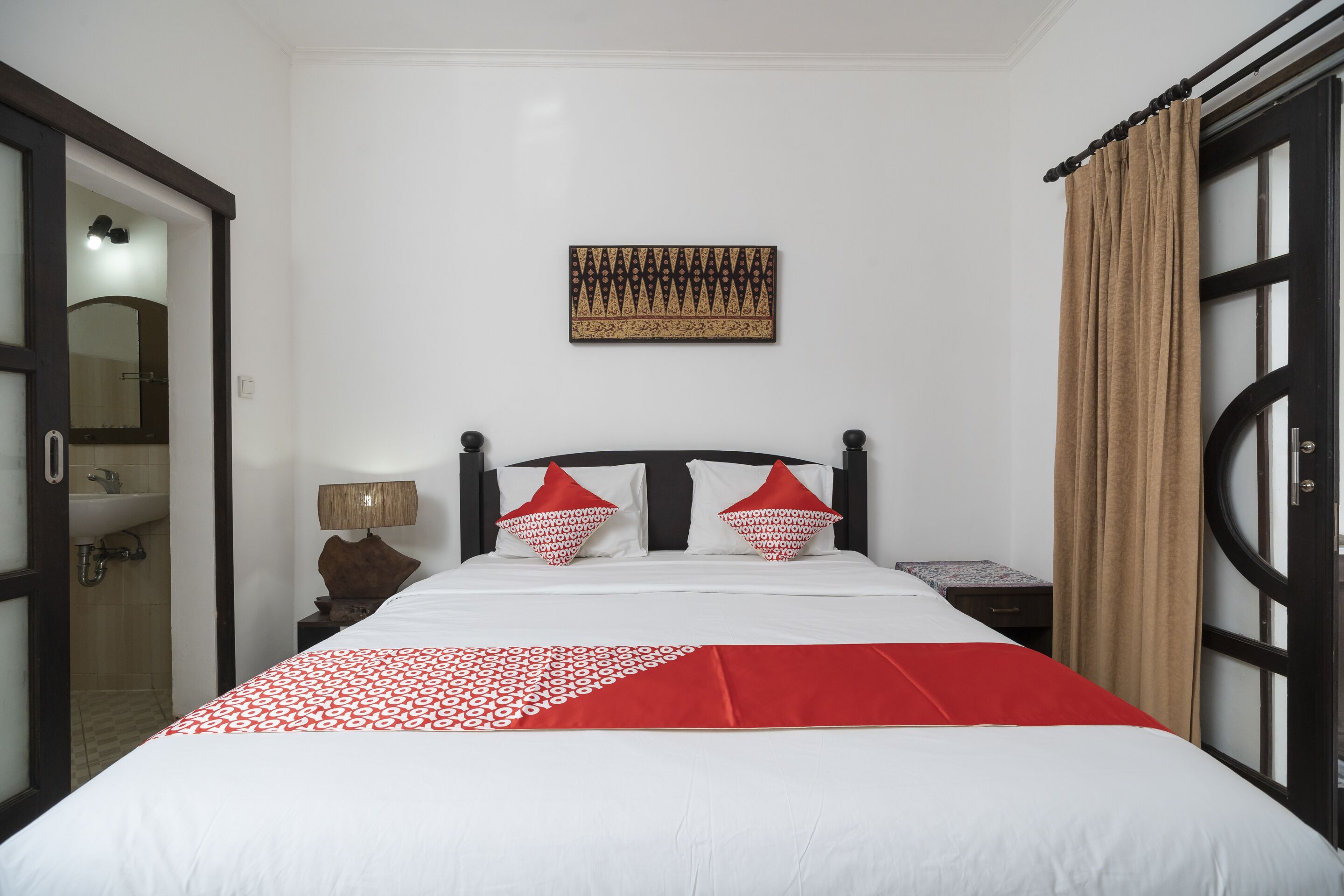 A hotel room with a bed, a nightstand, a lamp, a wall-mounted artwork, and a window with curtains.