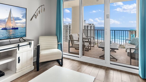 A balcony with a view of the ocean and buildings, a white chair, a table, and a TV displaying a sailboat. - ⭐By Beach in Seascape-Gulf Views-2 Pools + Kids Fountain-3BR Ariel Dunes I 1609 (Miramar Beach)