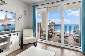 A balcony with a view of the ocean and buildings, a white chair, a table, and a TV displaying a sailboat.