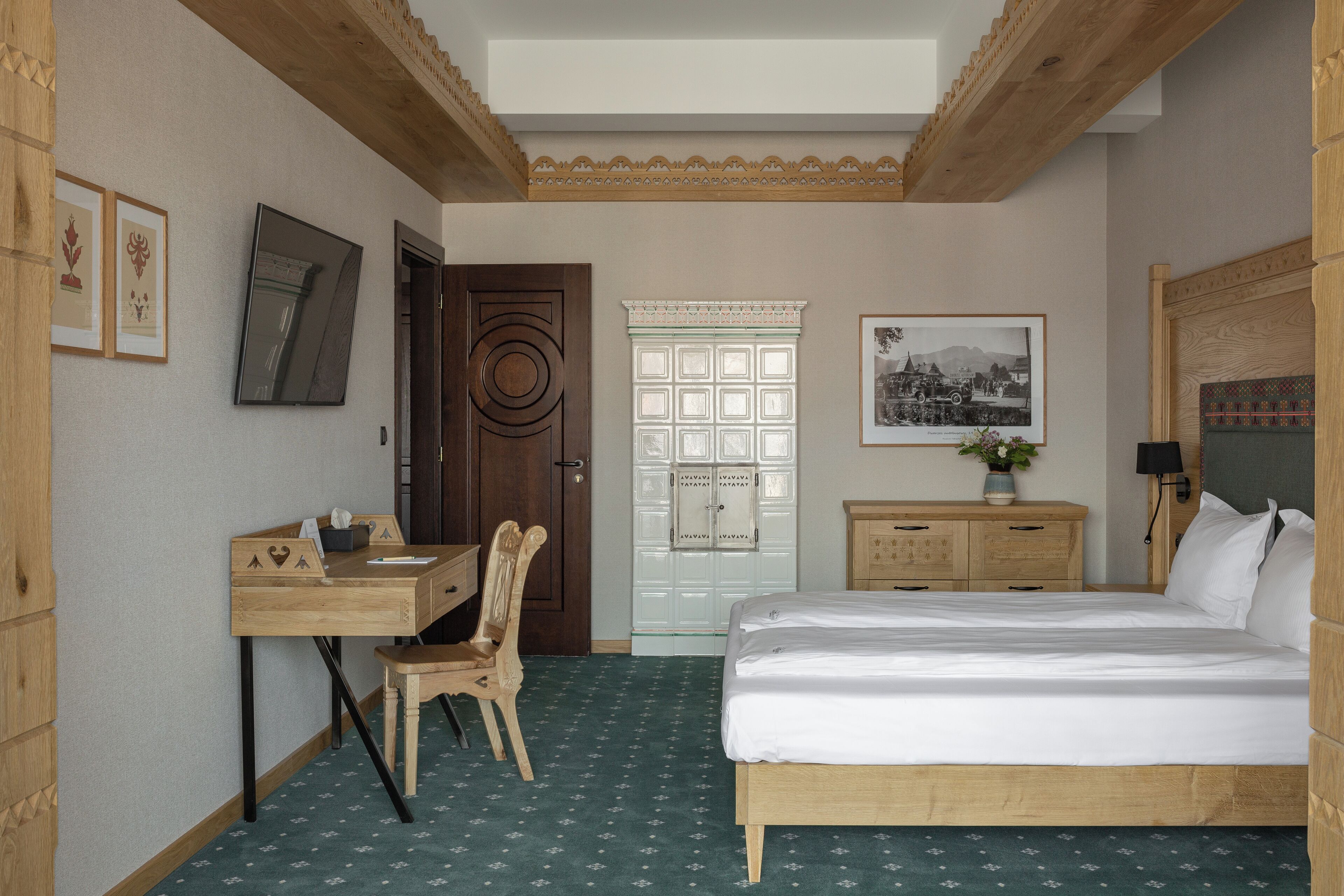 A bedroom with a wooden bed, a desk with a chair, a wardrobe, a door, and a framed picture on the wall.