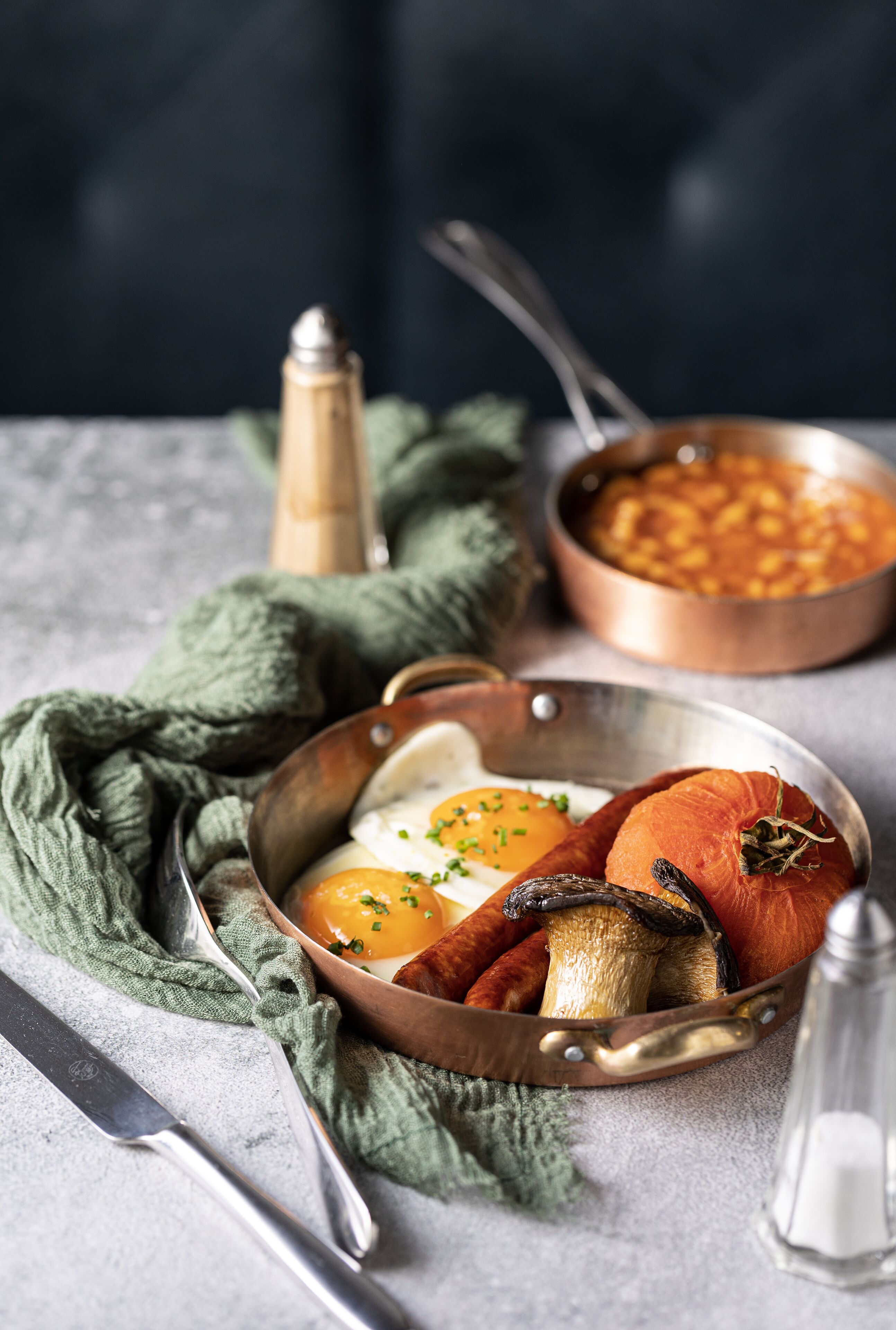A breakfast setting with eggs, mushrooms, and tomatoes in a pan, a bowl of beans, and a salt shaker on a table.