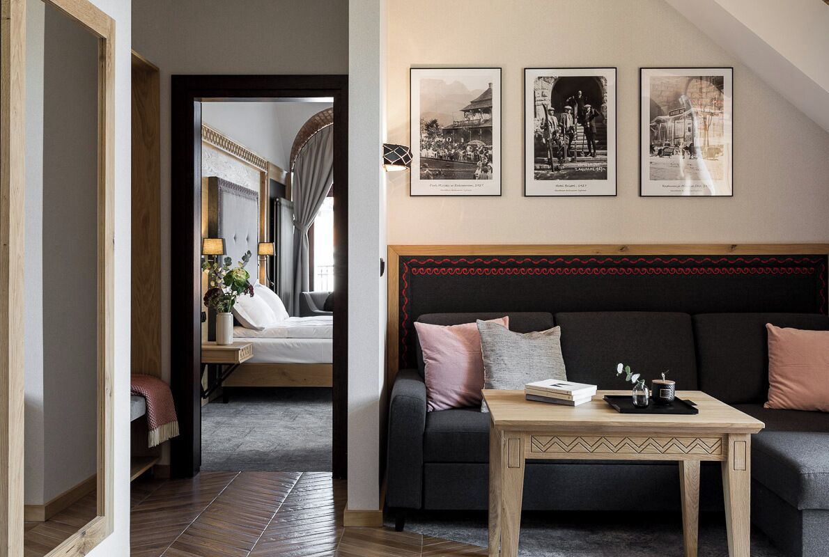 A modern hotel room with a dark sofa, a wooden coffee table, and a bed with a view of a balcony. There are three framed photographs on the wall.